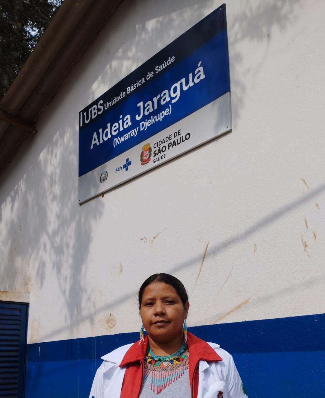 A foto mostra uma mulher jovem, de traços indígenas, cabelo preso e usando colar e brincos de miçanga coloridas e um jaleco branco; ela está junto a uma parede com uma placa onde se lê “UBS Aldeia Jaraguá Kwarãy Djekupé”, e possui também a logomarca do SUS e o brasão da Cidade de São Paulo 